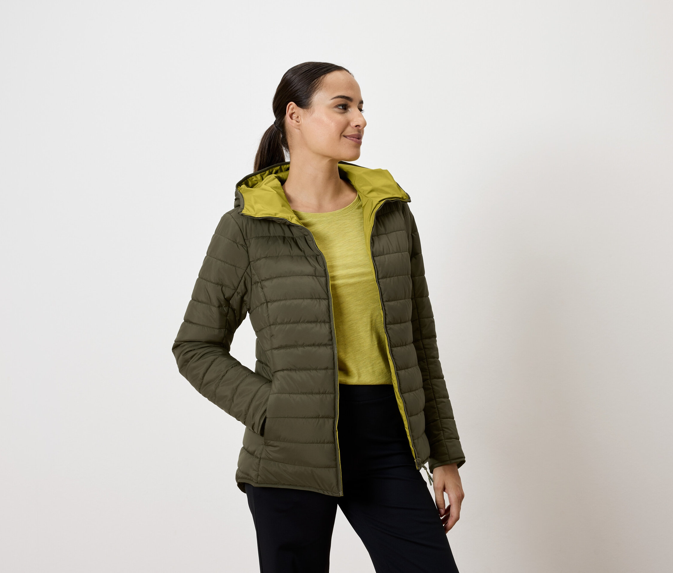 Frau trägt eine olivgrüne Steppjacke mit Kapuze, die Hände in den Taschen, und schaut nach rechts.