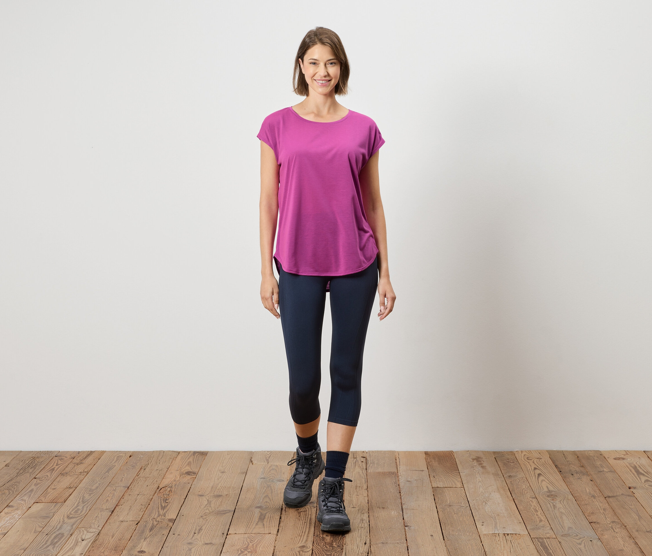 Eine Frau steht auf einem Holzboden vor einer weißen Wand. Sie trägt ein pinkfarbenes T-Shirt, blaue Leggings und graue Stiefel.