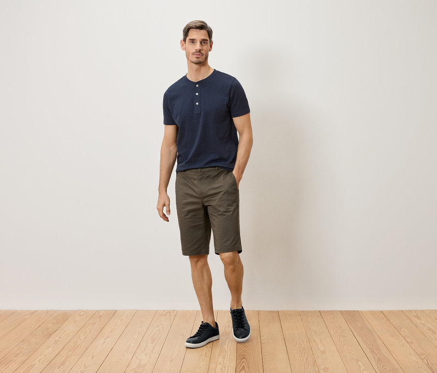 Ein Mann in marineblauem T-Shirt mit Knopfleiste, khakifarbener Shorts und marineblauen Sneakers steht vor einer weißen Wand.