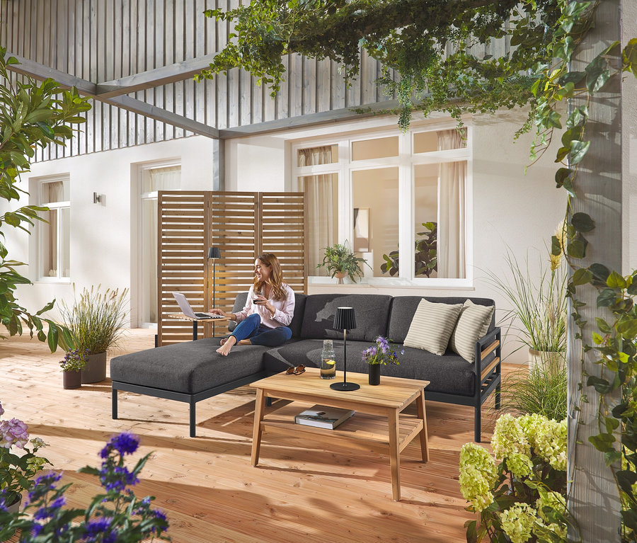 Eine Frau sitzt auf der Premium-Garten-Lounge-Ecke »Elin« mit einem flexiblen Paravent im Hintergrund. Eine schwarze LED-Akku-Outdoor-Steh- und Tischleuchte ist ebenfalls zu sehen, sowie ein Garten-Couchtisch.