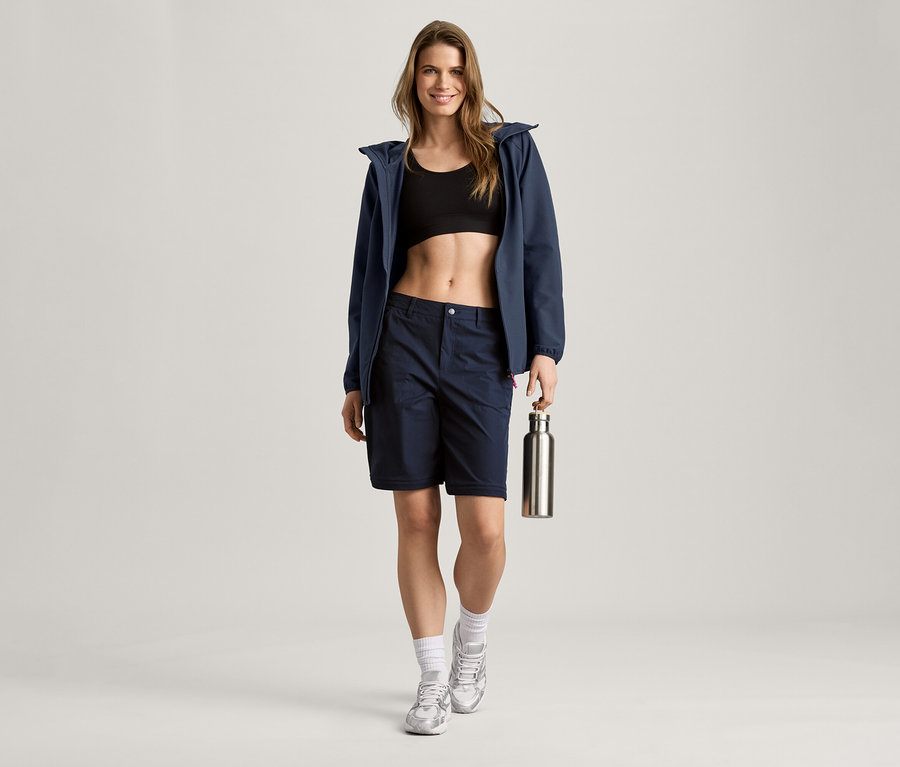 Frau mit navy Softshelljacke, Shorts und Sport-BH hält eine silberne Flasche.