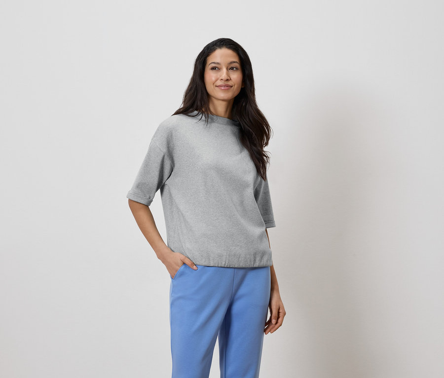 Frau mit dunklen Haaren trägt ein graues Oberteil und blaue Hosen, die Hand in der Hosentasche.