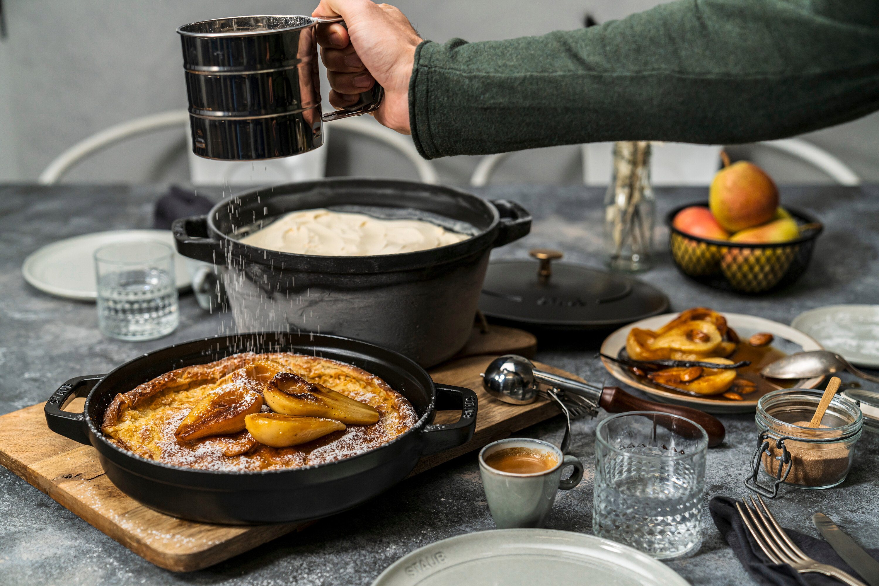 Eine Hand streut Puderzucker auf einen Dutch Baby mit Birnen; daneben ein Staub Gusseisen-Kochtopf-Set mit Teig.