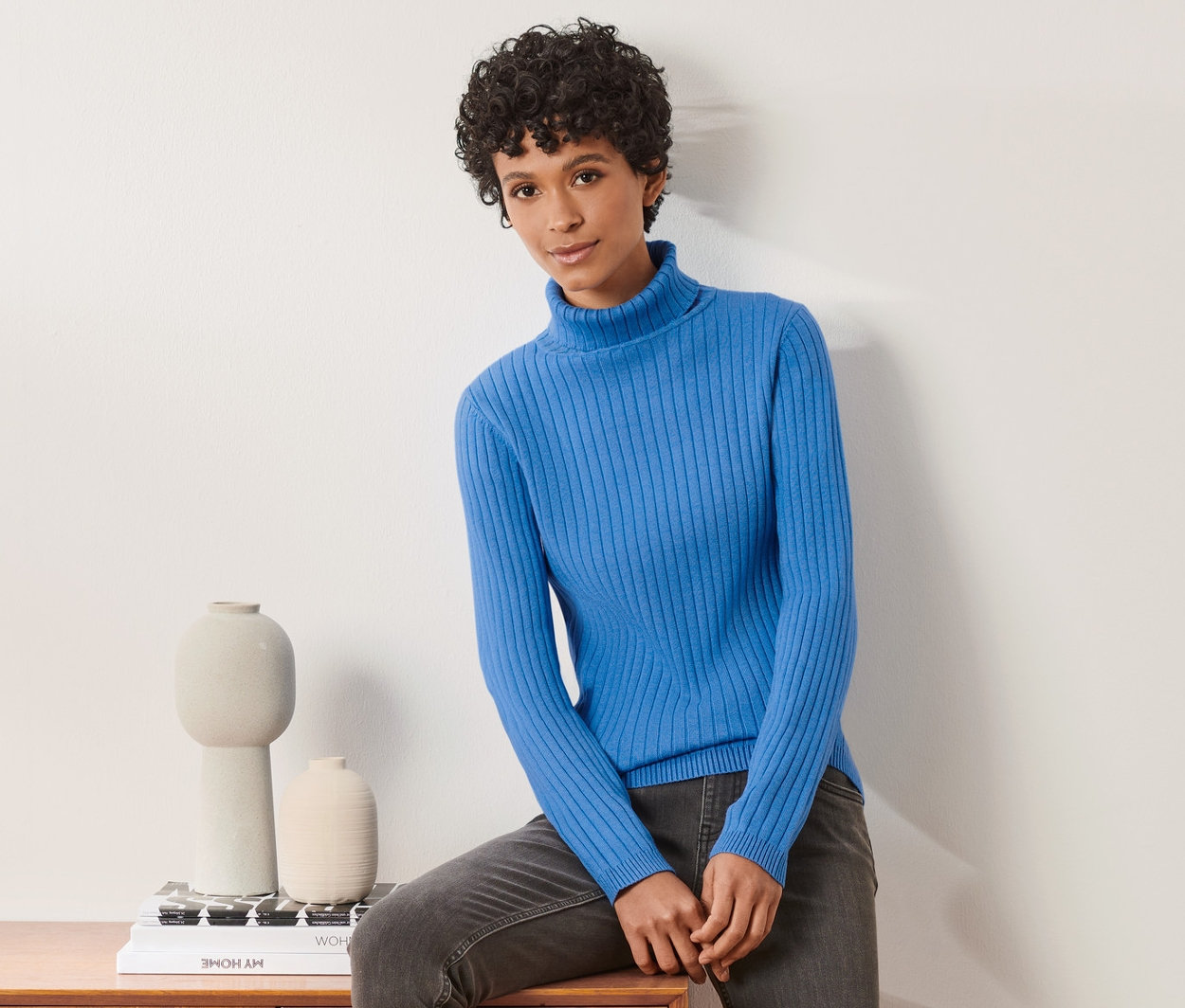 Frau sitzt auf einer Holzkommode und trägt einen gerippten Rollkragenpullover.