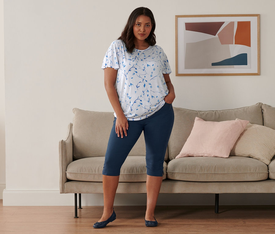 Eine Frau steht in einem Raum und trägt ein weißes Blusenshirt mit Flügelärmeln, eine dunkelblaue Bengalin-Hose in 3/4-Länge und Leder-Ballerinas.