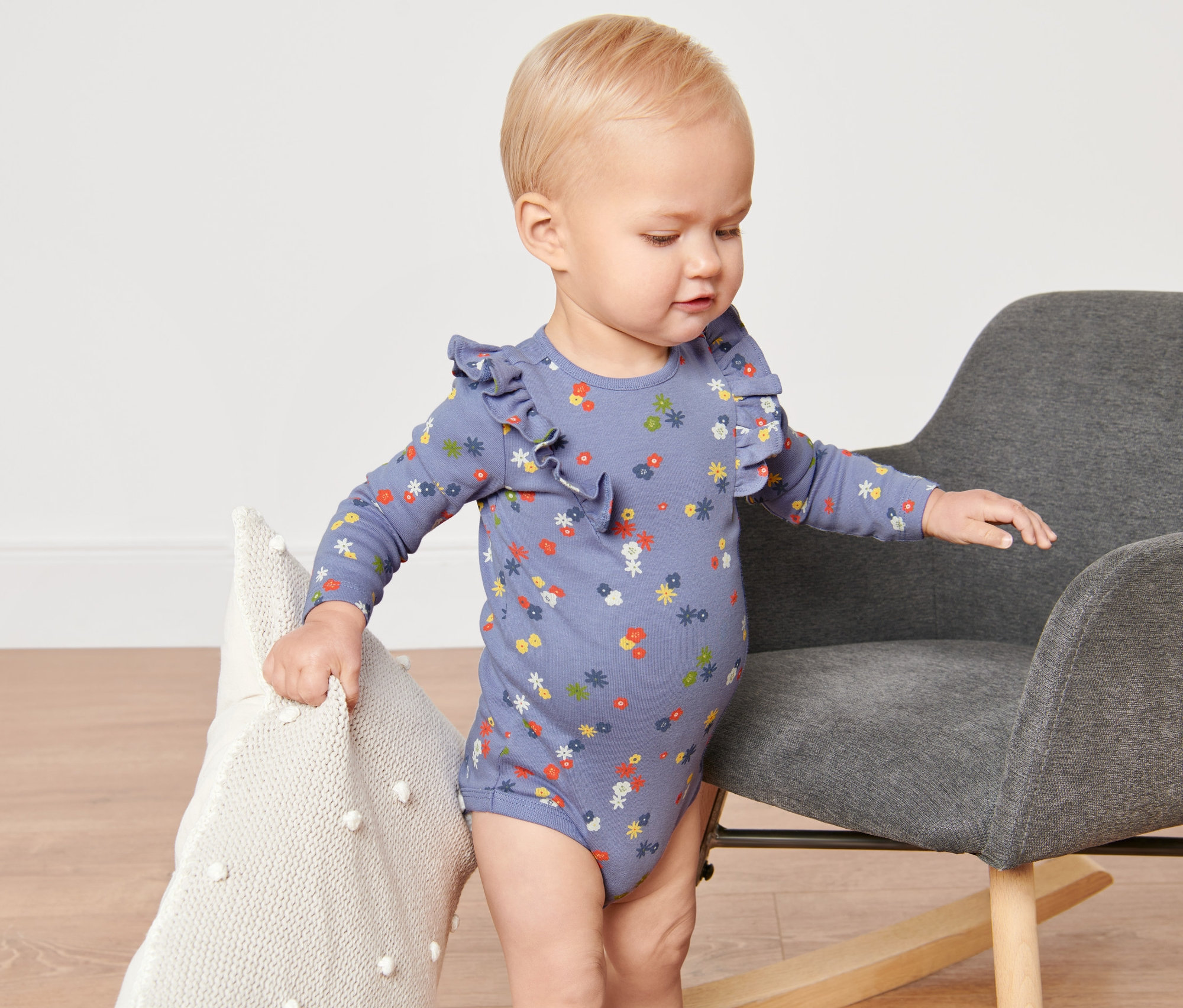 Kleinkind steht neben einem grauen Sessel und hält ein weisses Kissen. Es trägt einen Baby-Body mit Blumenmuster.