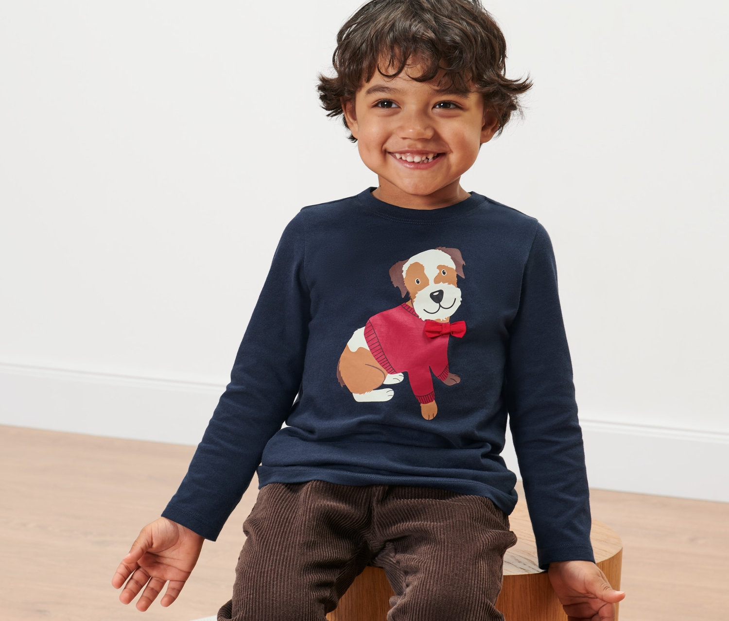 Ein lächelnder Junge sitzt und trägt ein Kleinkinder-Langarmshirt mit einem Hundemotiv.