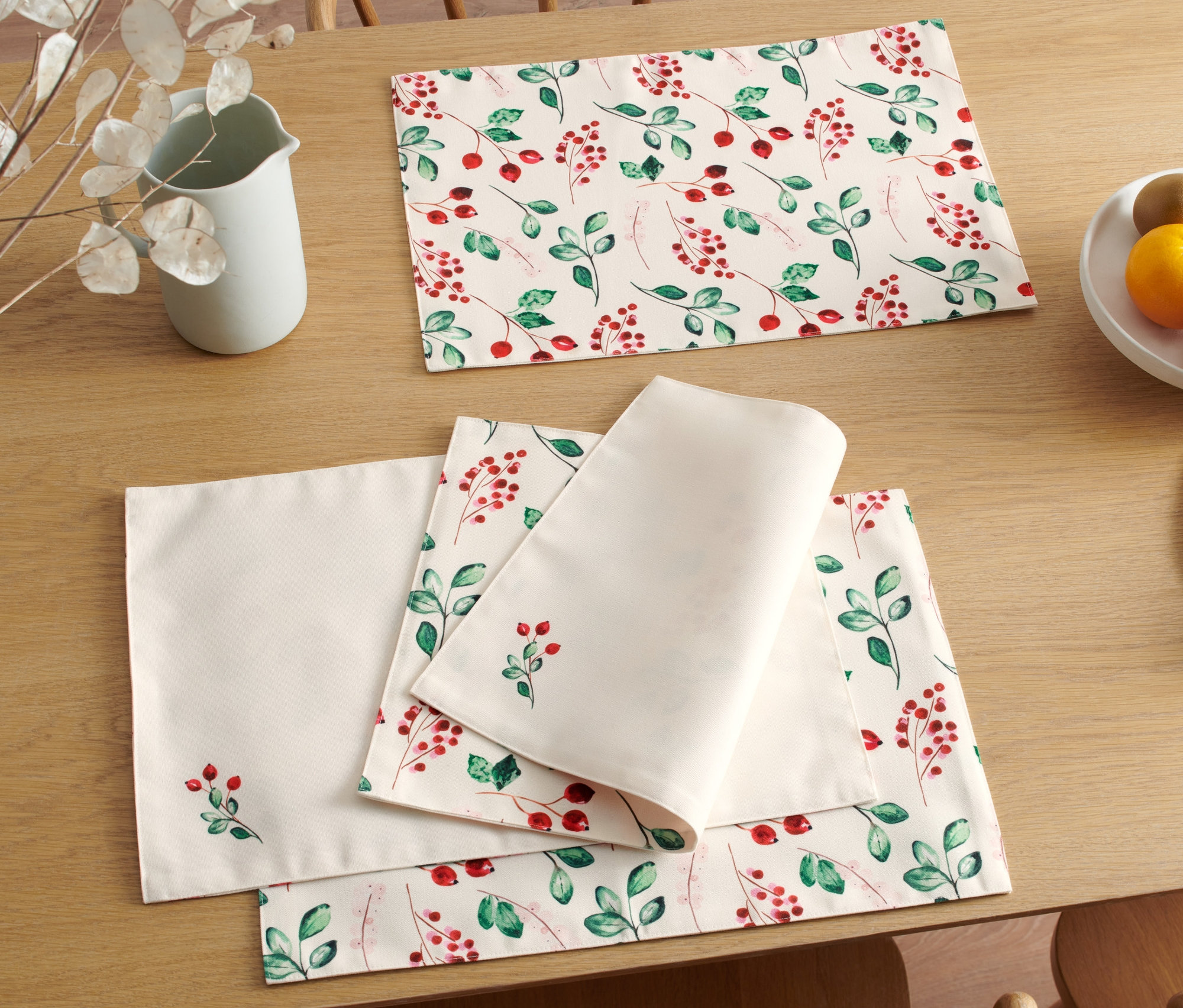 Auf einem Holztisch liegen 4 Wende-Platzsets mit Weihnachtsmotiv, ein Krug, Obst und Brot.