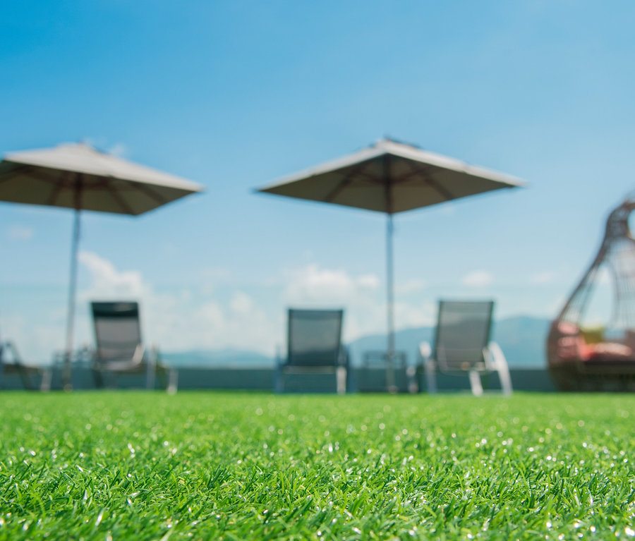 Nahaufnahme von Primaflor Kunstrasen »WOODLAND« mit Liegestühlen und Sonnenschirmen im Hintergrund.