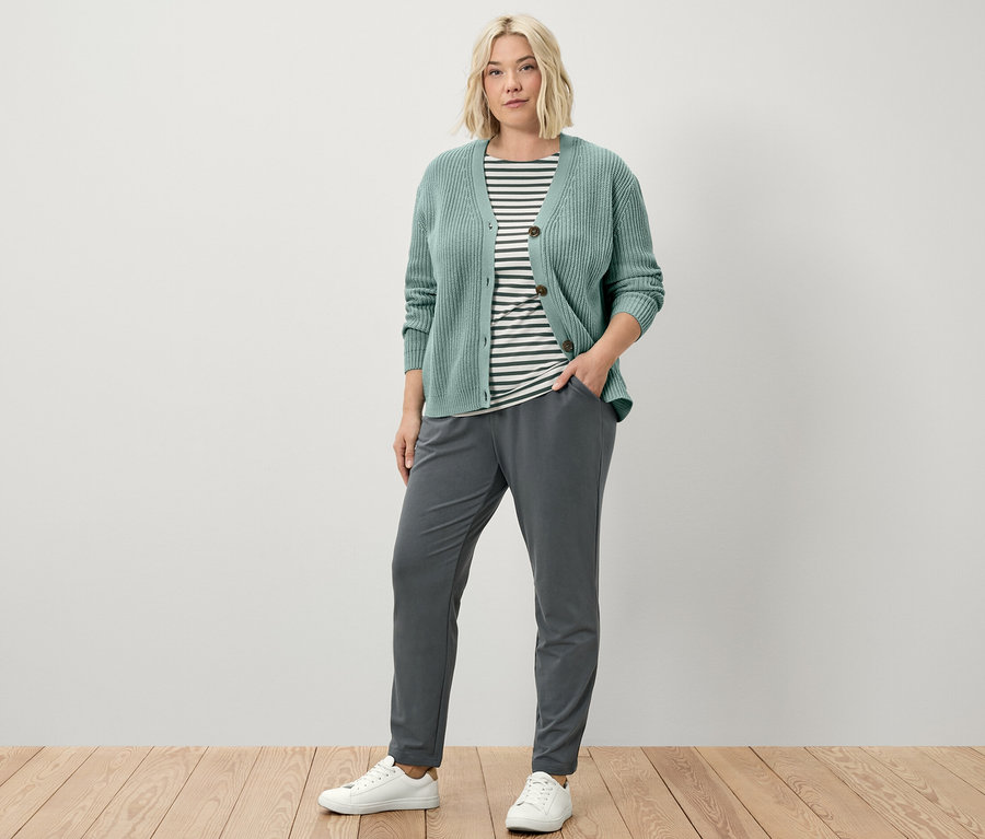 Eine Frau steht und trägt Joggpants, einen grob gestrickten Cardigan in Salbei und ein grün gestreiftes Halbarmshirt.