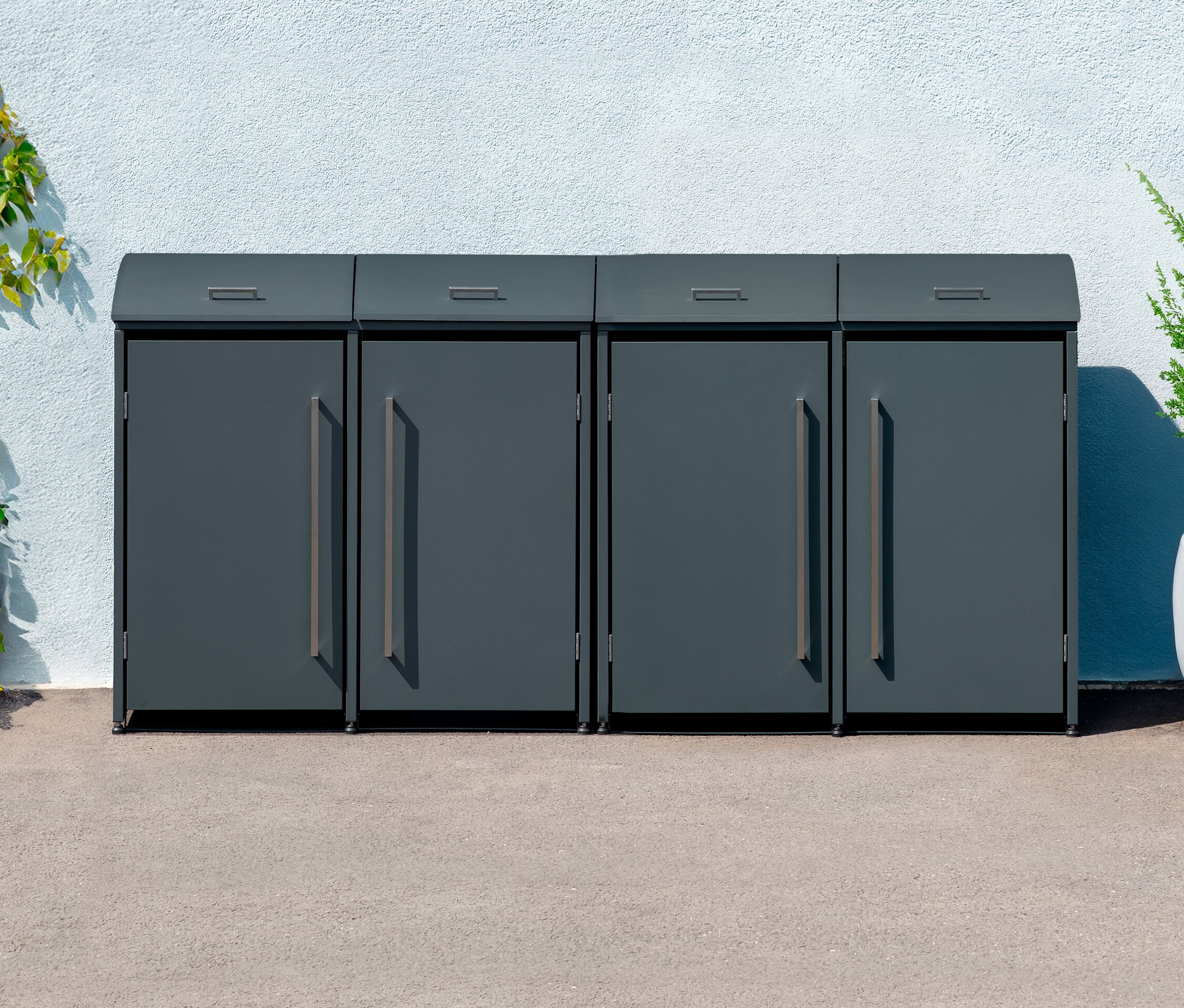 Vier anthrazitfarbene Mülltonnenboxen stehen nebeneinander vor einer weißen Wand, daneben ein Strauch.