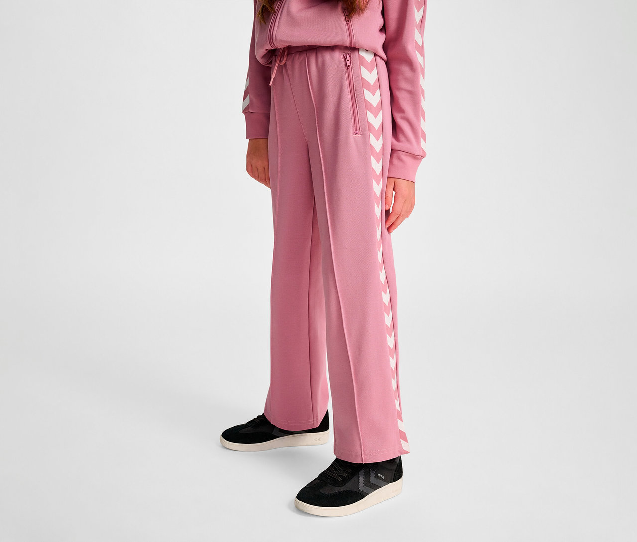 Person trägt eine HUMMEL HMLJR Archive Wideleg Poly Pants. Sie ist rosa mit weißen Winkelstreifen an den Seiten.