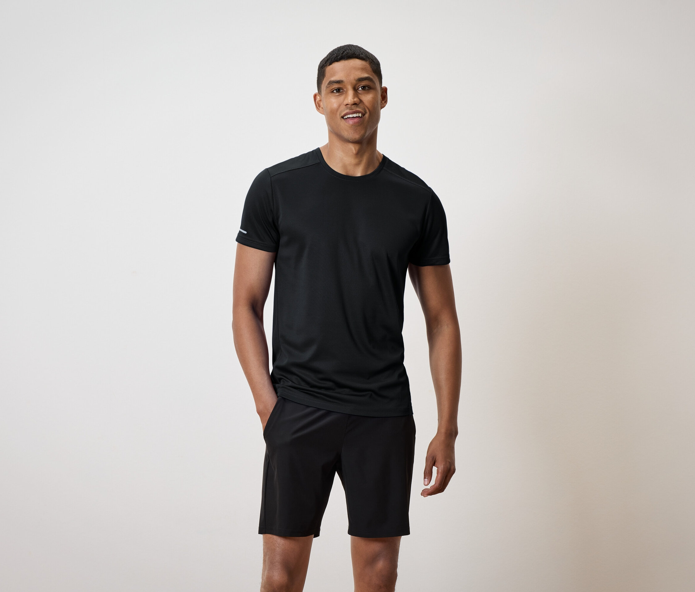 Ein lächelnder Mann steht mit den Händen in den Taschen seiner schwarzen Shorts und trägt ein schwarzes T-Shirt.