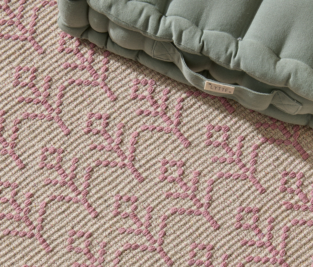 Nahaufnahme eines beigen Teppichs mit rosafarbenem geometrischen Muster und einem grünen Sitzkissen mit 'LYTTE'-Etikett.