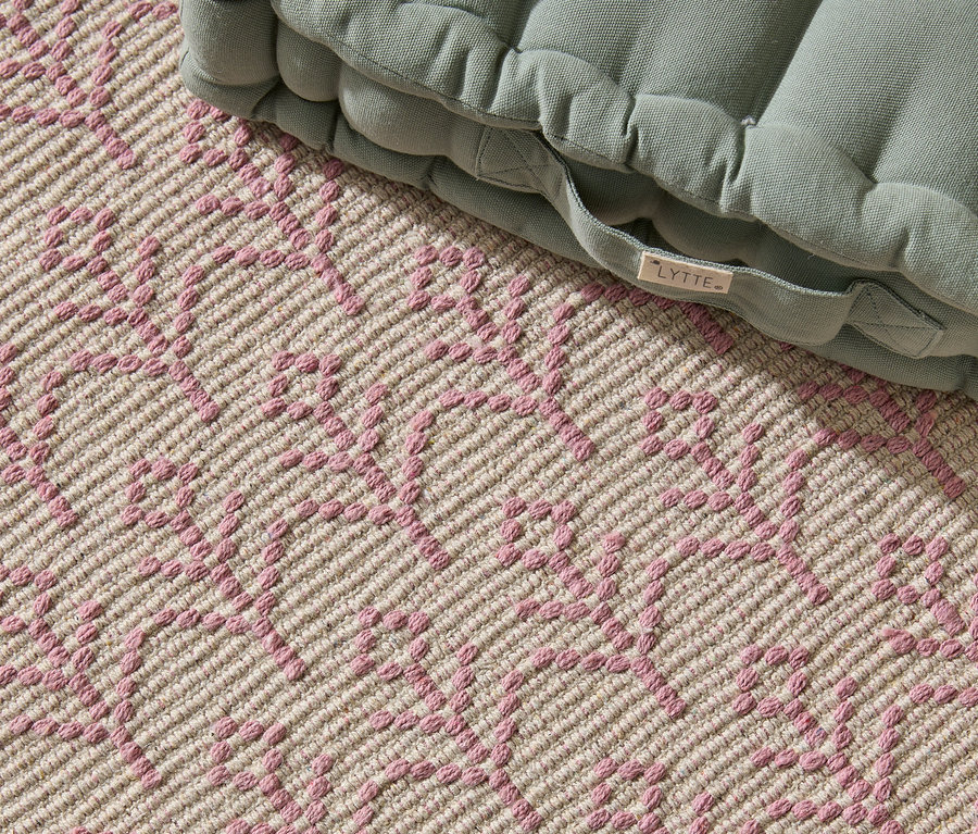 Nahaufnahme eines beigen Teppichs mit rosafarbenem geometrischen Muster und einem grünen Sitzkissen mit 'LYTTE'-Etikett.