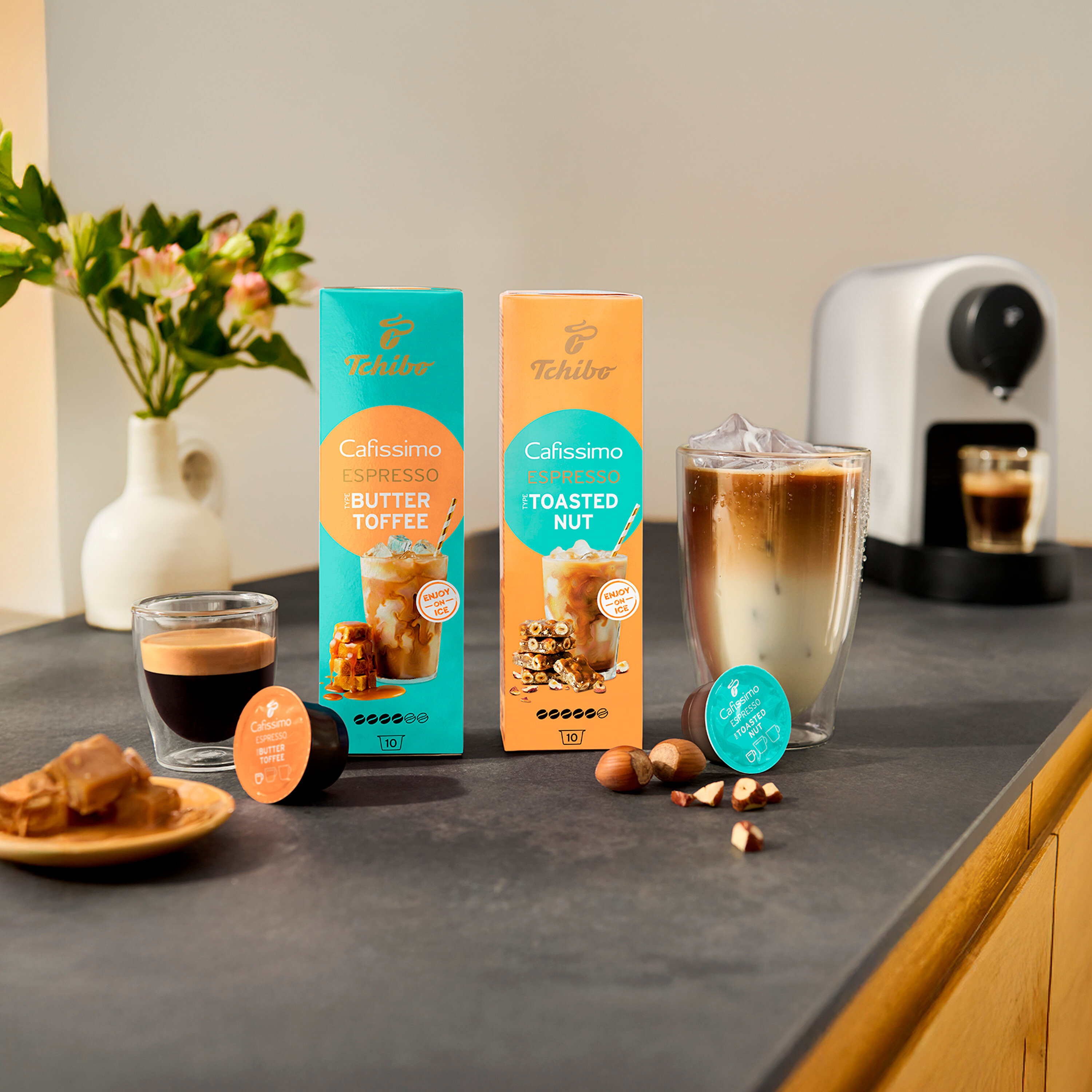 Auf einer Küchentheke stehen Tchibo Cafissimo Kapselpackungen Espresso Butter Toffee und Toasted Nut neben passenden Getränken und Zutaten.