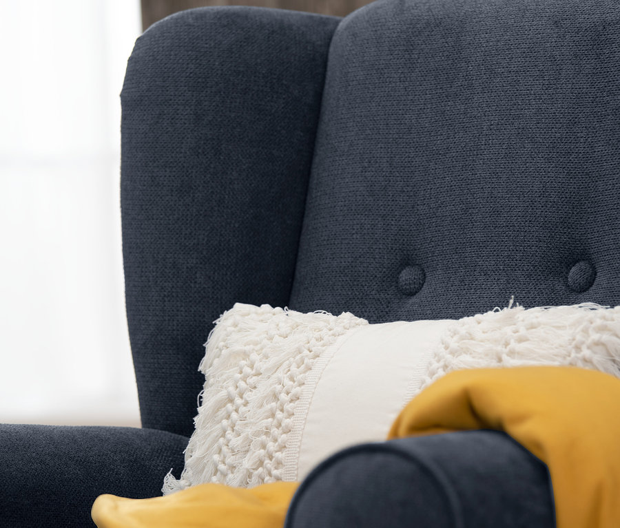 Detailansicht eines dunkelblauen Ohrensessels "Anton" mit einem weißen Kissen und einer gelben Decke.