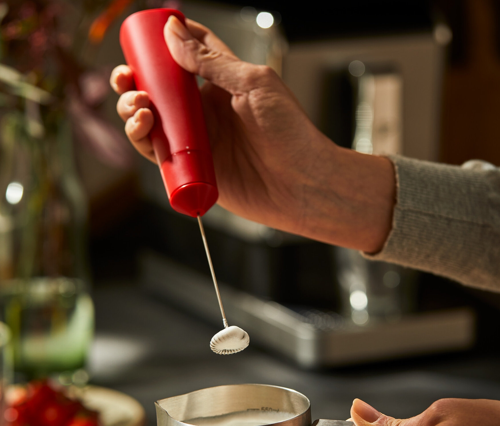 Frau schäumt Milch in einem Edelstahlkrug mit einem roten Batterie-Milchaufschäumer auf.