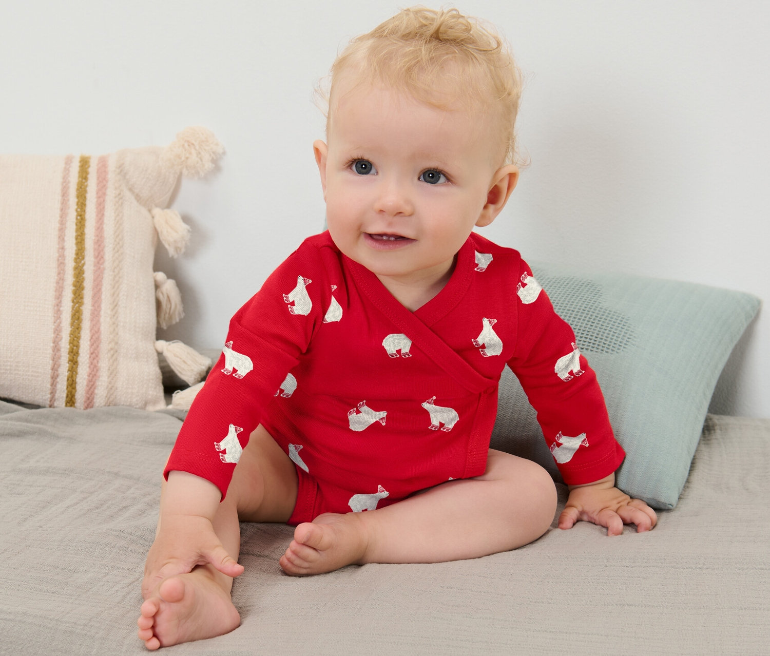 Ein blondes Baby Mädchen trägt einen roten Langarmbody mit Eisbärmuster und sitzt auf einer grauen Decke.