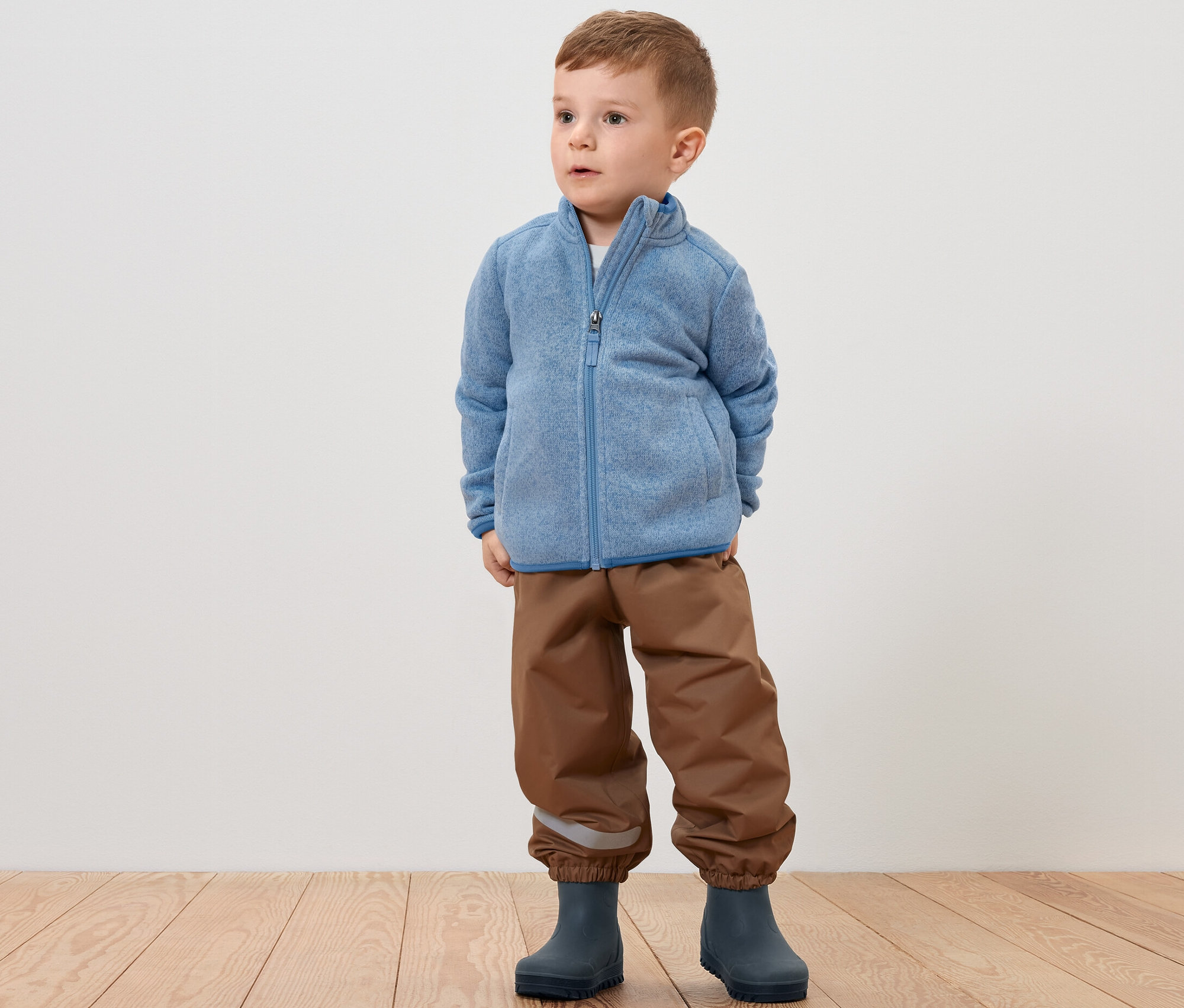 Ein Junge steht und trägt eine Kinder-Strickfleecejacke, braune Hosen und graue Stiefel.