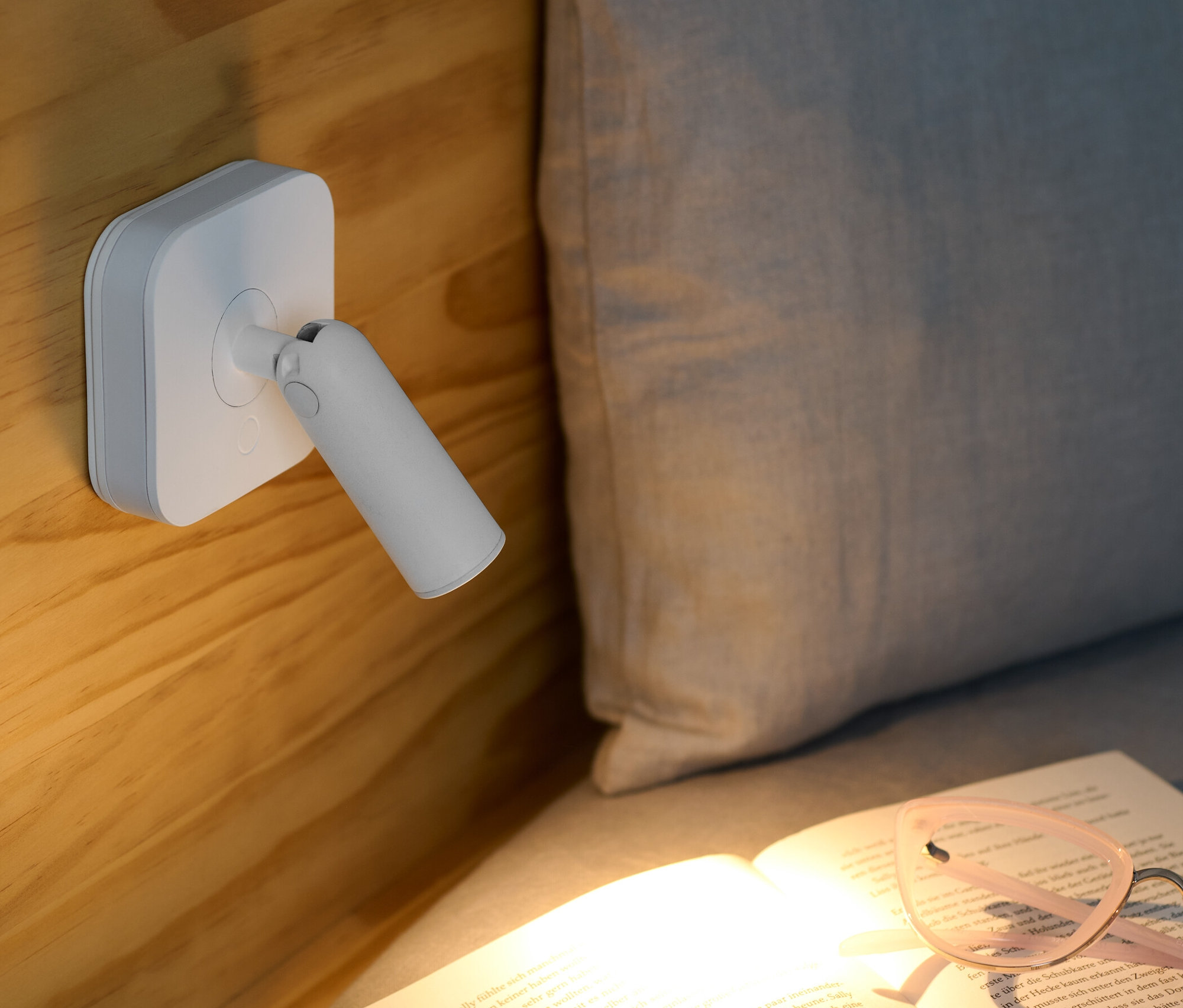 LED-Nachtlicht mit Spot ist an einem Holzbett befestigt und beleuchtet ein aufgeschlagenes Buch mit Brille.