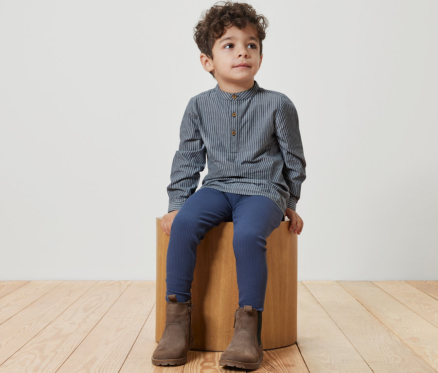 Junge sitzt auf einem Holzhocker und trägt ein Kinderhemd, blaue Leggings und braune Stiefel.