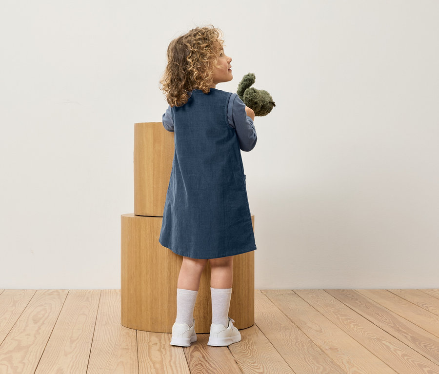 Ein Mädchen in einem blauen Kleid hält ein Stofftier und steht neben Holzblöcken.