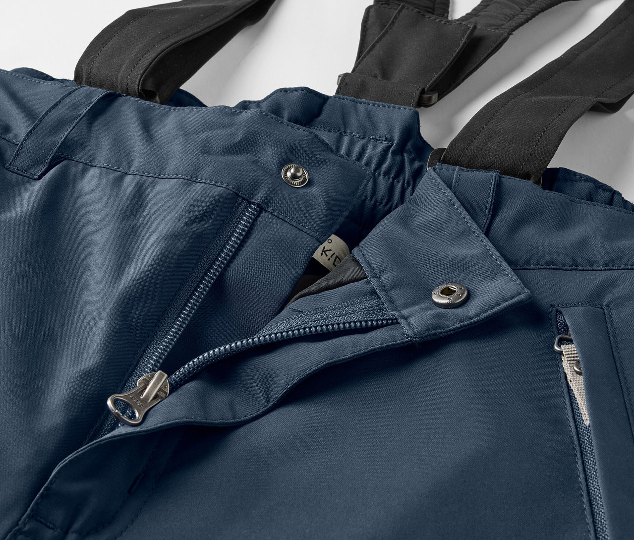 Detailaufnahme einer blauen Hose mit schwarzen Hosenträgern, offenem Reißverschluss und silbernen Knöpfen.