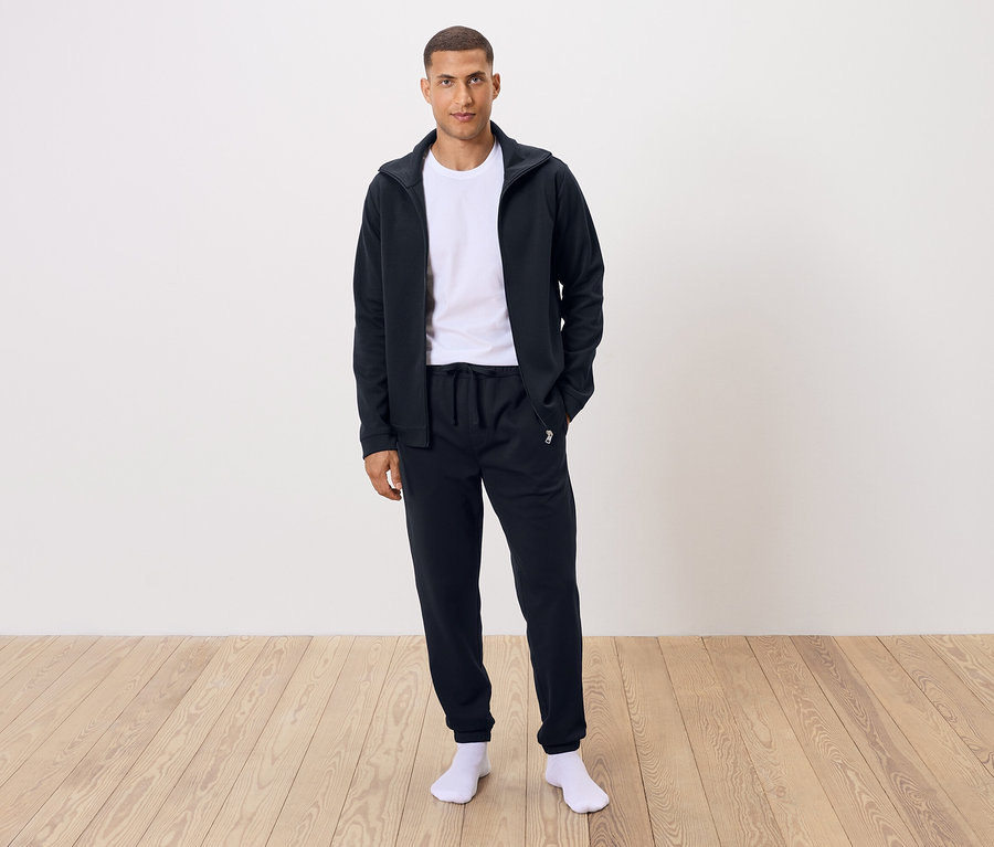 Mann steht in weißem T-Shirt, schwarzer Jogginghose und schwarzer Reißverschlussjacke.