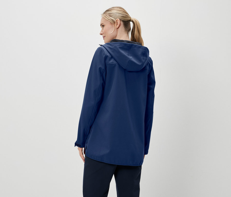 Rückansicht einer Frau mit blonden, zum Pferdeschwanz gebundenen Haaren, die eine dunkelblaue Jacke mit Kapuze und dunkle Hosen trägt.