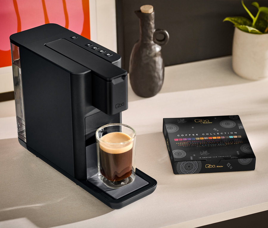 Eine Qbo ICONIC Dark Chrome Kaffeemaschine mit einer Tasse Kaffee daneben und eine Qbo Collection - 16 Kapseln Packung stehen auf einem Tisch.