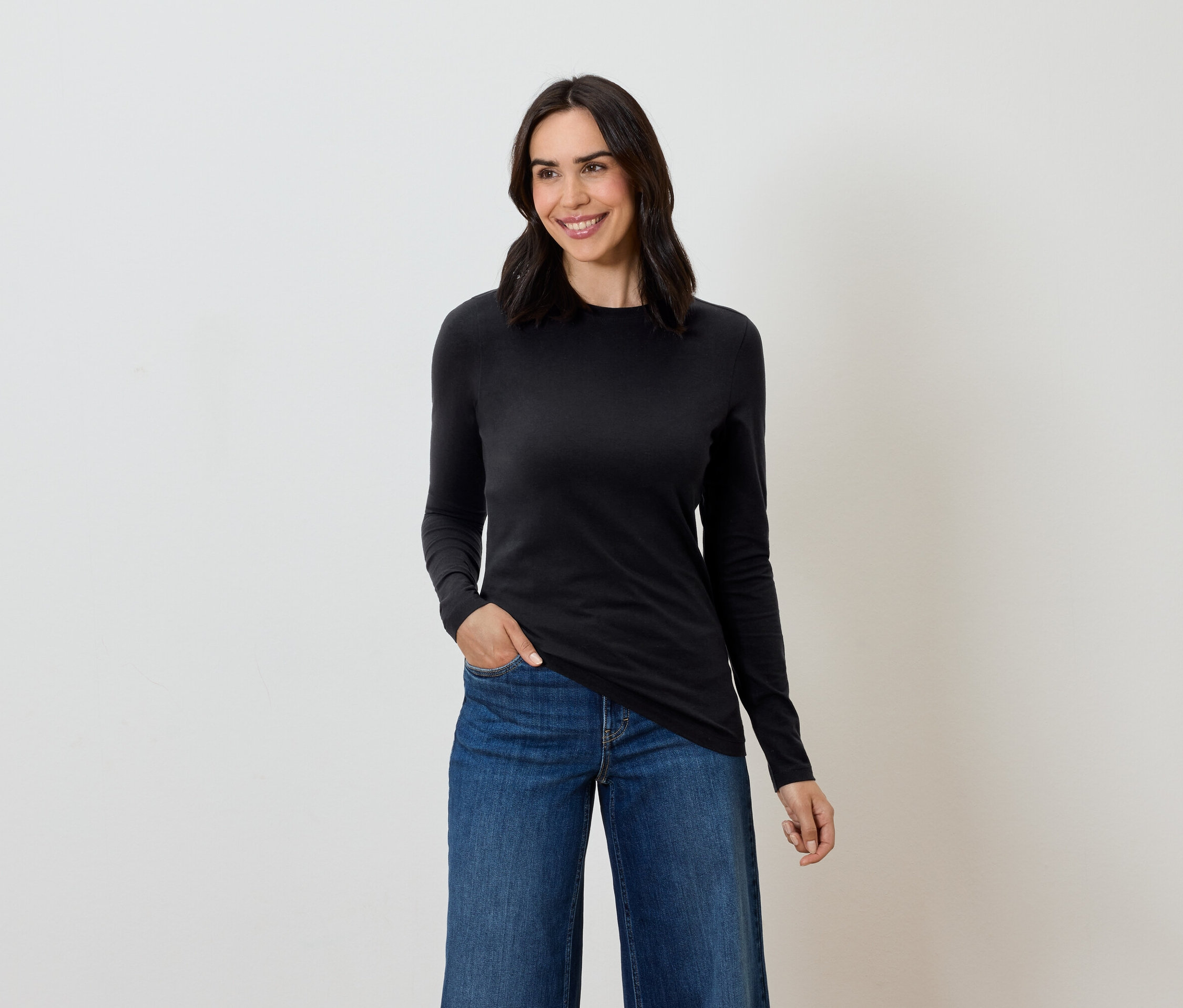 Frau mit braunen Haaren posiert in einem schwarzen Langarmshirt und blauen Jeans.