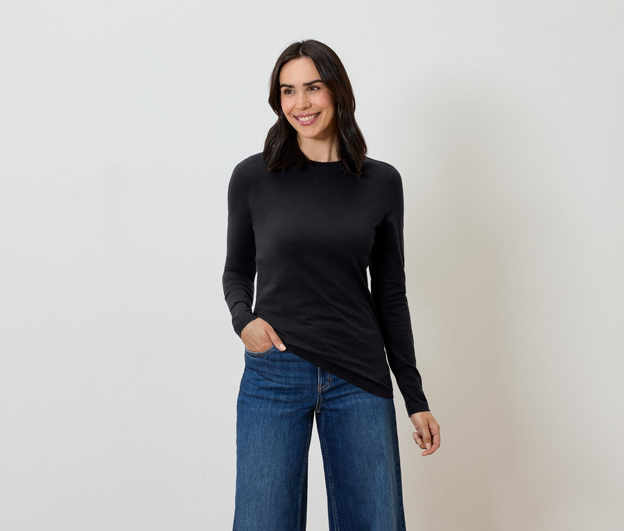 Frau mit braunen Haaren posiert in einem schwarzen Langarmshirt und blauen Jeans.