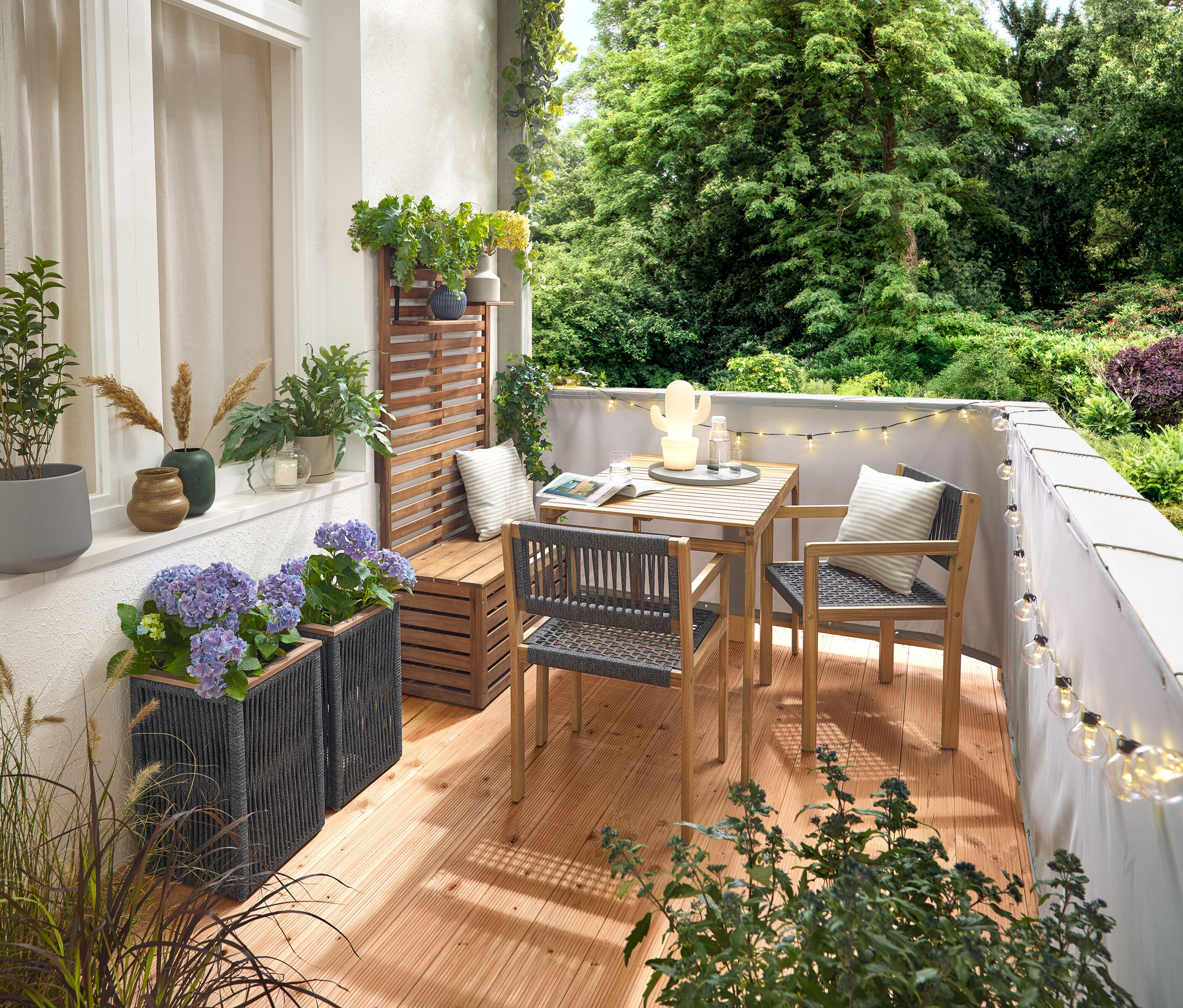 Einrichtung eines Balkons mit einem Balkon-Set mit Textilgeflecht, einem grauen Balkon Sicht- und Windschutz, einer LED-Outdoor-Lichterkette mit Netzadapter, einem Pflanztopf mit Textilgeflecht, einer LED-Outdoor-Akku-Tischleuchte in Kaktusform und 2 Outdoor-Kissen mit Leinen-Haptik.
