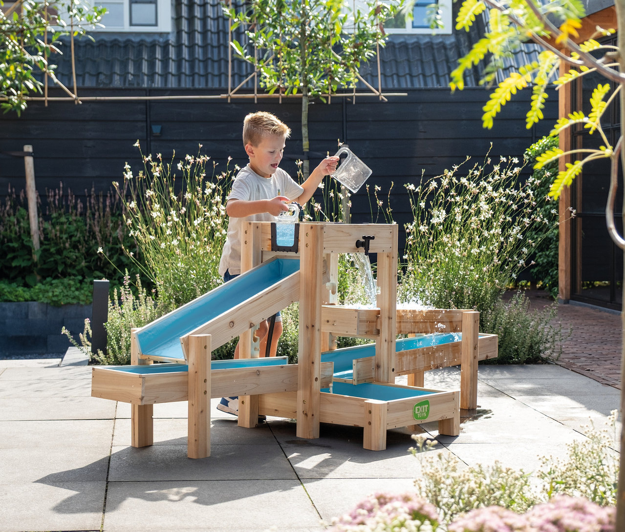 Ein Junge gießt Wasser aus einem Krug in eine hölzerne Wasserspielbahn mit blauen Rinnen im Garten.