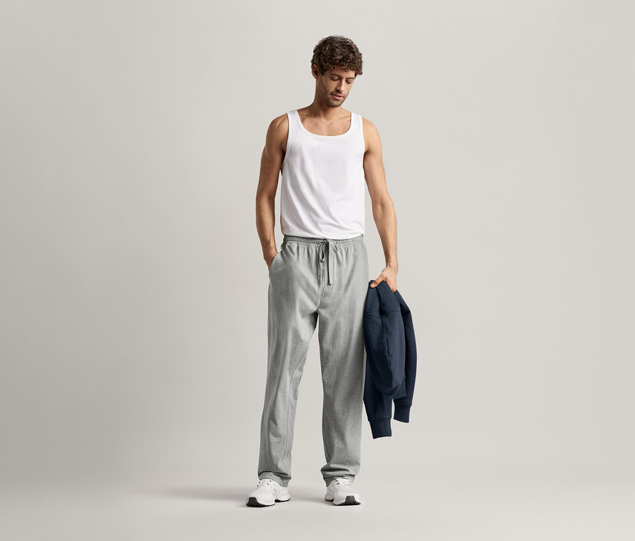 Mann mit weißem Tanktop und grauer Relaxhose, mit einer Hand in der Hosentasche und einer marineblauen Jacke über dem Arm.