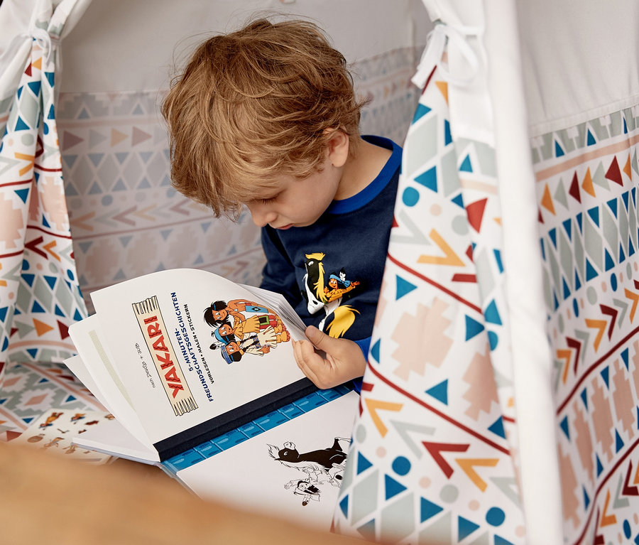Ein Junge liest ein Yakari-Buch im Pinolino Zelt »Tipi Naira«. Er trägt einen Yakari-Pyjama und neben ihm liegen ein Yakari-Plüschtier-Set und 3 Paar Yakari-Socken.