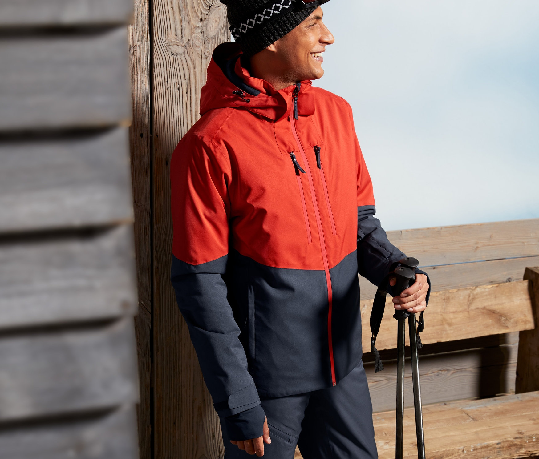 Mann in rot-blauer Skijacke, an eine Holzwand gelehnt, hält Skistöcke.