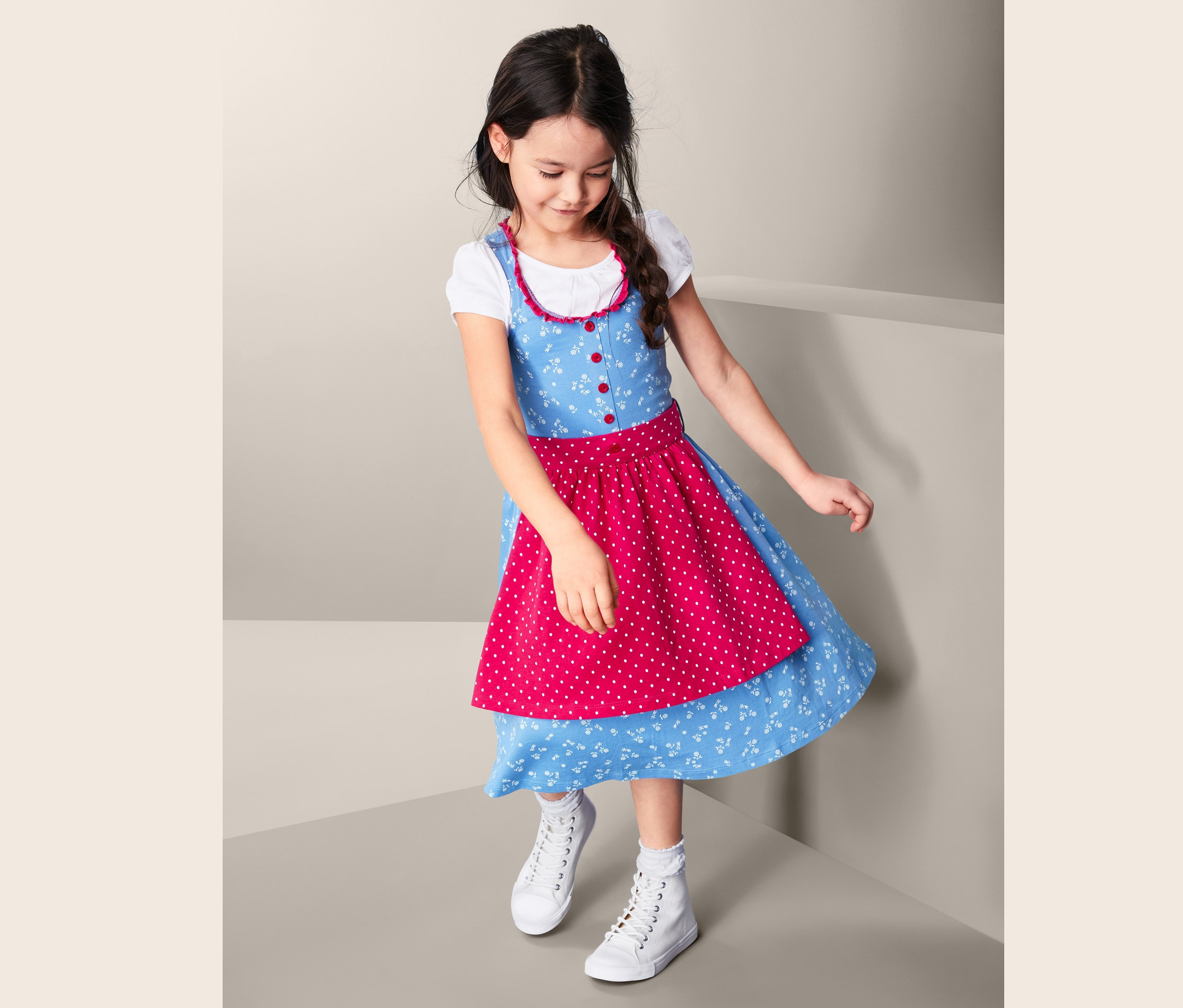 Mädchen trägt ein blaues Jersey-Dirndl mit roter Schürze mit weißen Punkten und weißen Turnschuhen.