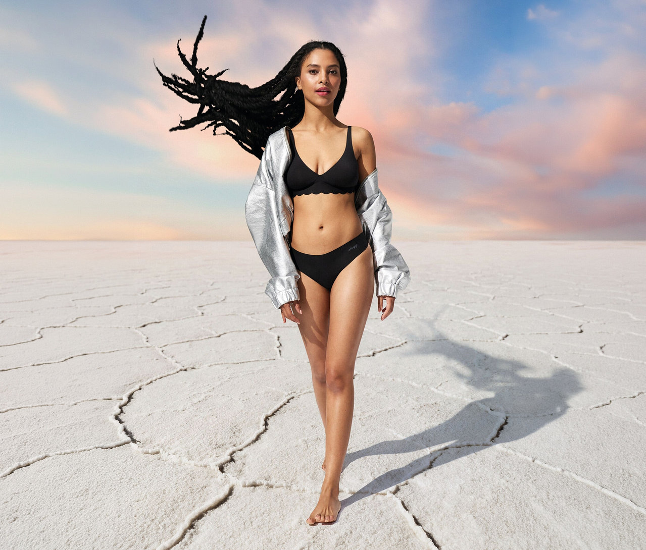 Frau mit silberner Jacke und schwarzem Sloggi ZERO Microfibre 2.0 Soft BH auf weißem Grund.