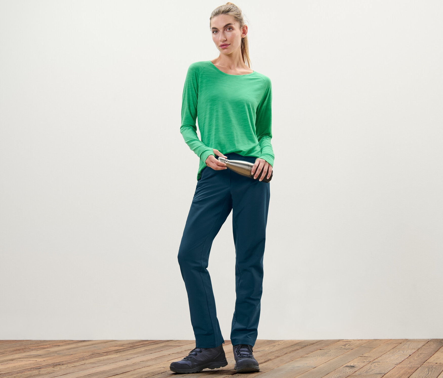 Frau im grünen Shirt hält eine silberne Flasche und trägt eine Softshellhose.