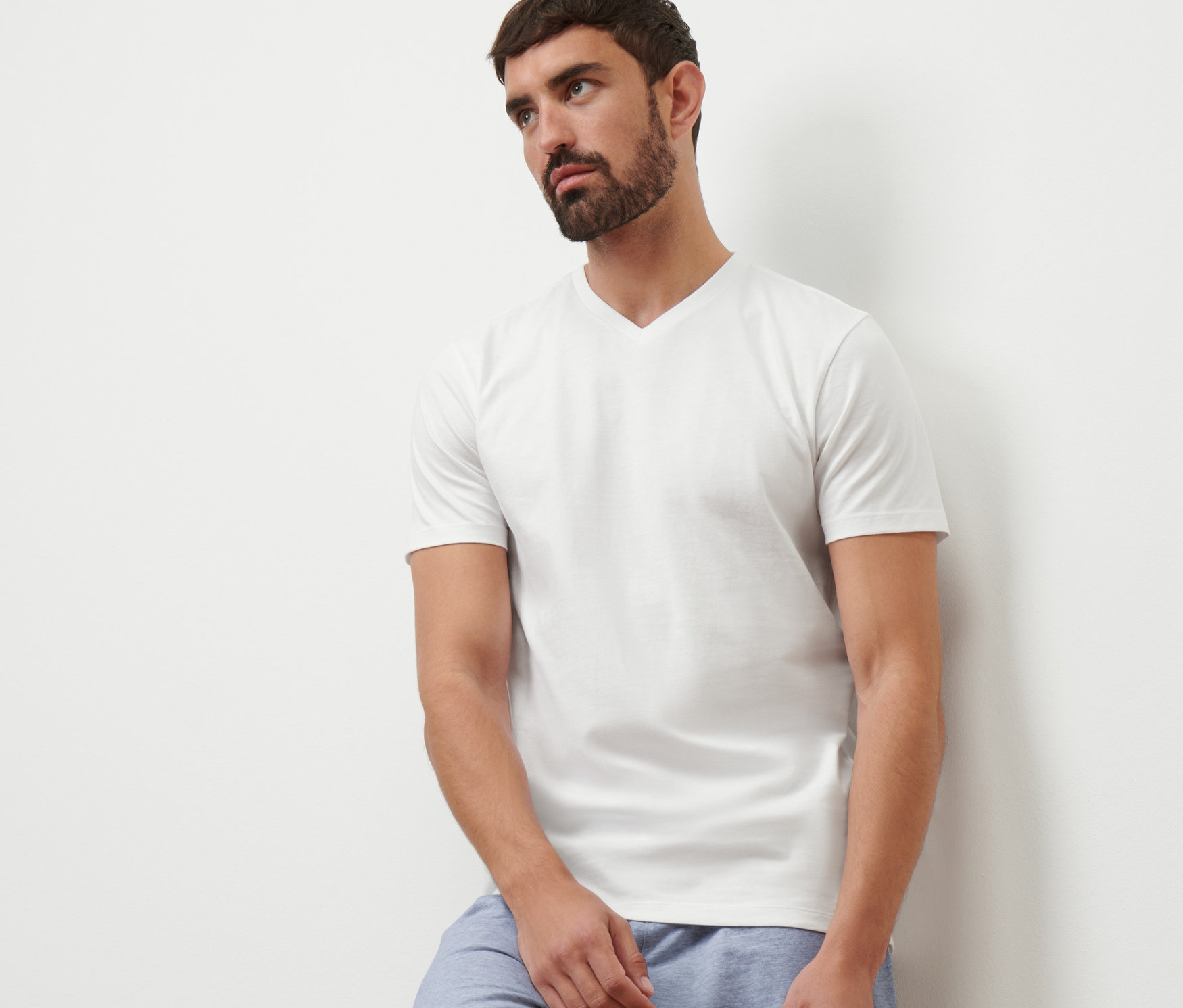 Mann lehnt an einer Wand, trägt ein weißes V-Ausschnitt T-Shirt und blaue Shorts.