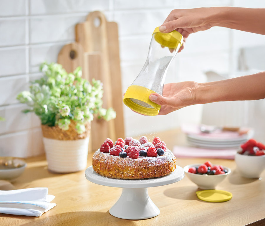 Eine Person streut Puderzucker mit einem gelben Streuer auf einen Kuchen mit Beeren.