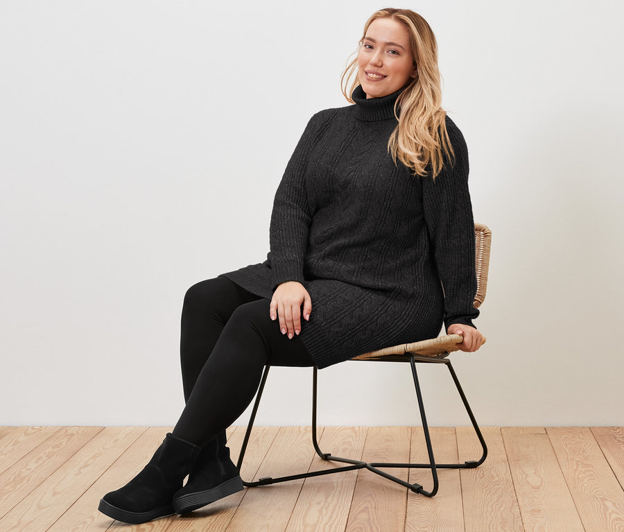 Eine Frau sitzt auf einem Stuhl und trägt einen langen, schwarzen Strickpullover mit Zopfmuster.