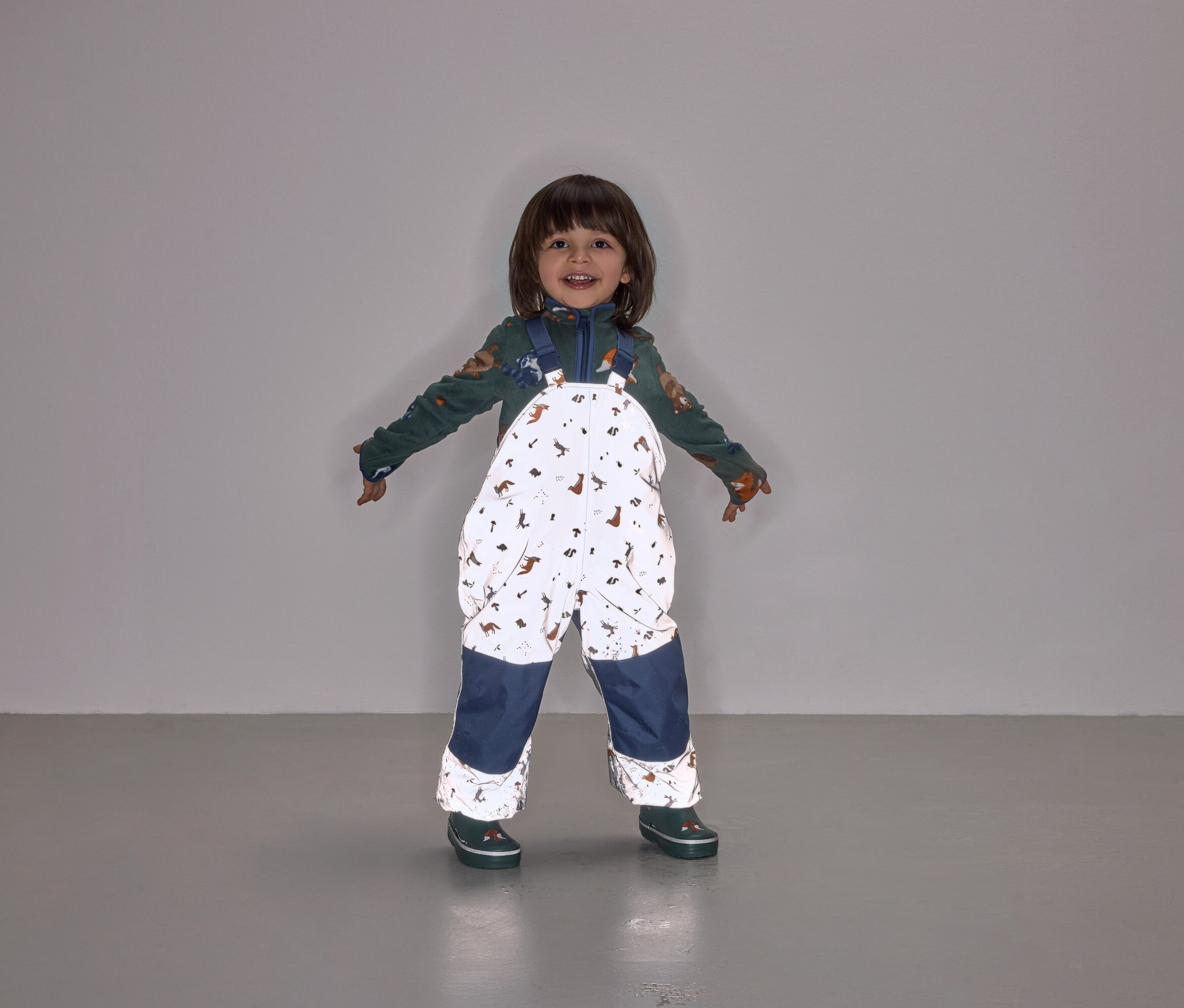 Ein lächelndes Kind steht mit ausgestreckten Armen und trägt eine Latzhose mit Tiermuster und marineblauen Hosenbeinen.