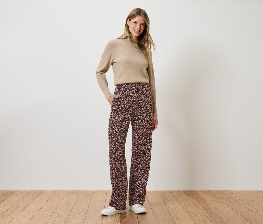 Frau steht mit Händen in den Hosentaschen, trägt eine braune, geblümte Hose und einen beigen Pullover.