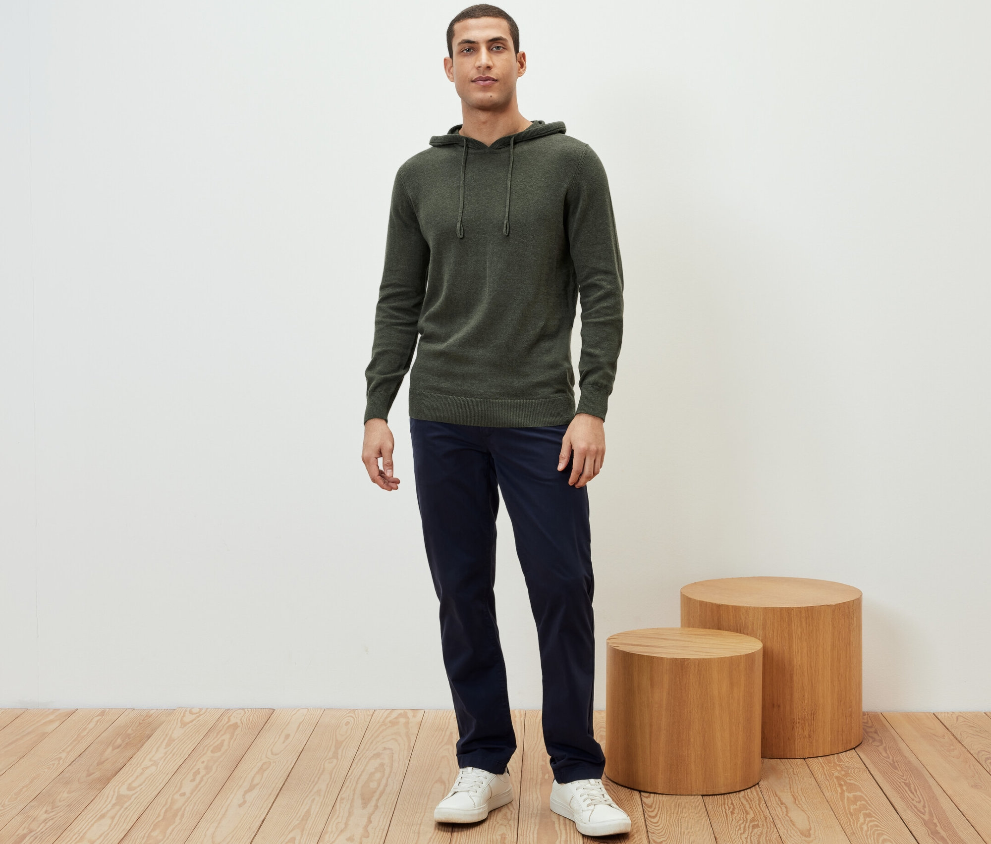 Mann steht in Strick-Hoodie, marineblauer Hose und weißen Turnschuhen.