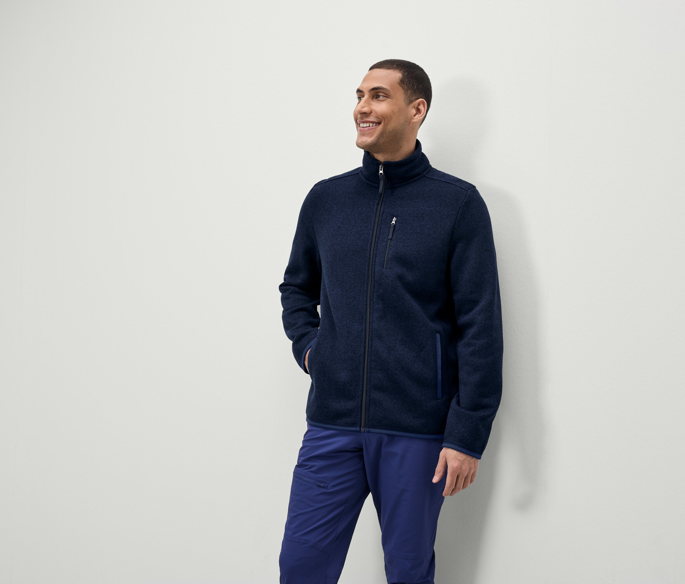 Ein Mann lächelt und schaut zur Seite, er trägt eine blaue Fleecejacke mit Reißverschluss und blaue Hosen.