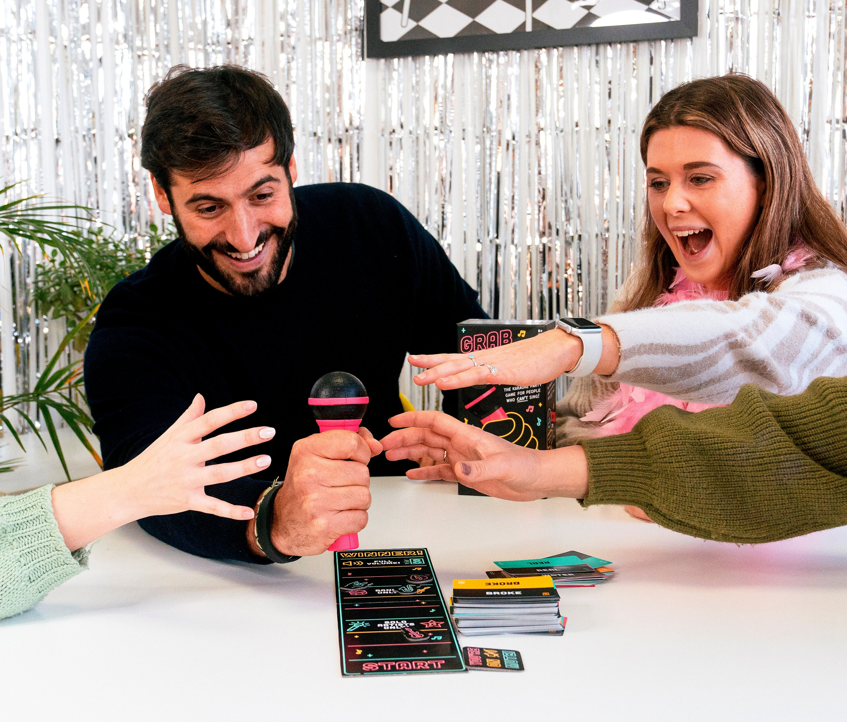 Mann hält ein pinkes Mikrofon in der Hand, während eine Frau und zwei weitere Personen um das Spiel »Grab the Mic« herumstehen.