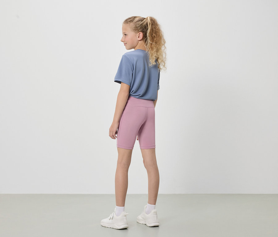 Mädchen mit blonden Haaren, von hinten gesehen, trägt ein blaues T-Shirt, kurze, rosafarbene Leggings und weiße Turnschuhe vor einem weißen Hintergrund.